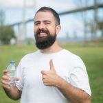 Cheerful man with a beard giving thumbs up while holding a water bottle in a park setting in Portugal.